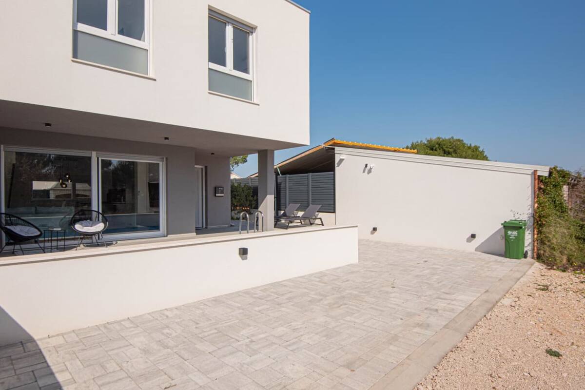 A modern two-story house with a patio, a covered outdoor seating area, and a visible trash bin.