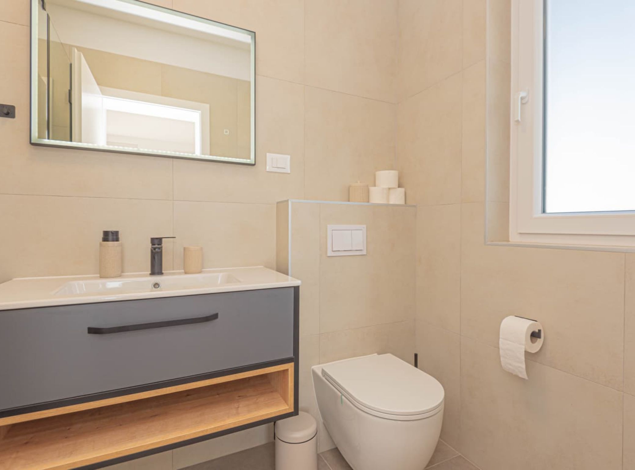 A modern bathroom with a sink, mirror, toilet, and a window.