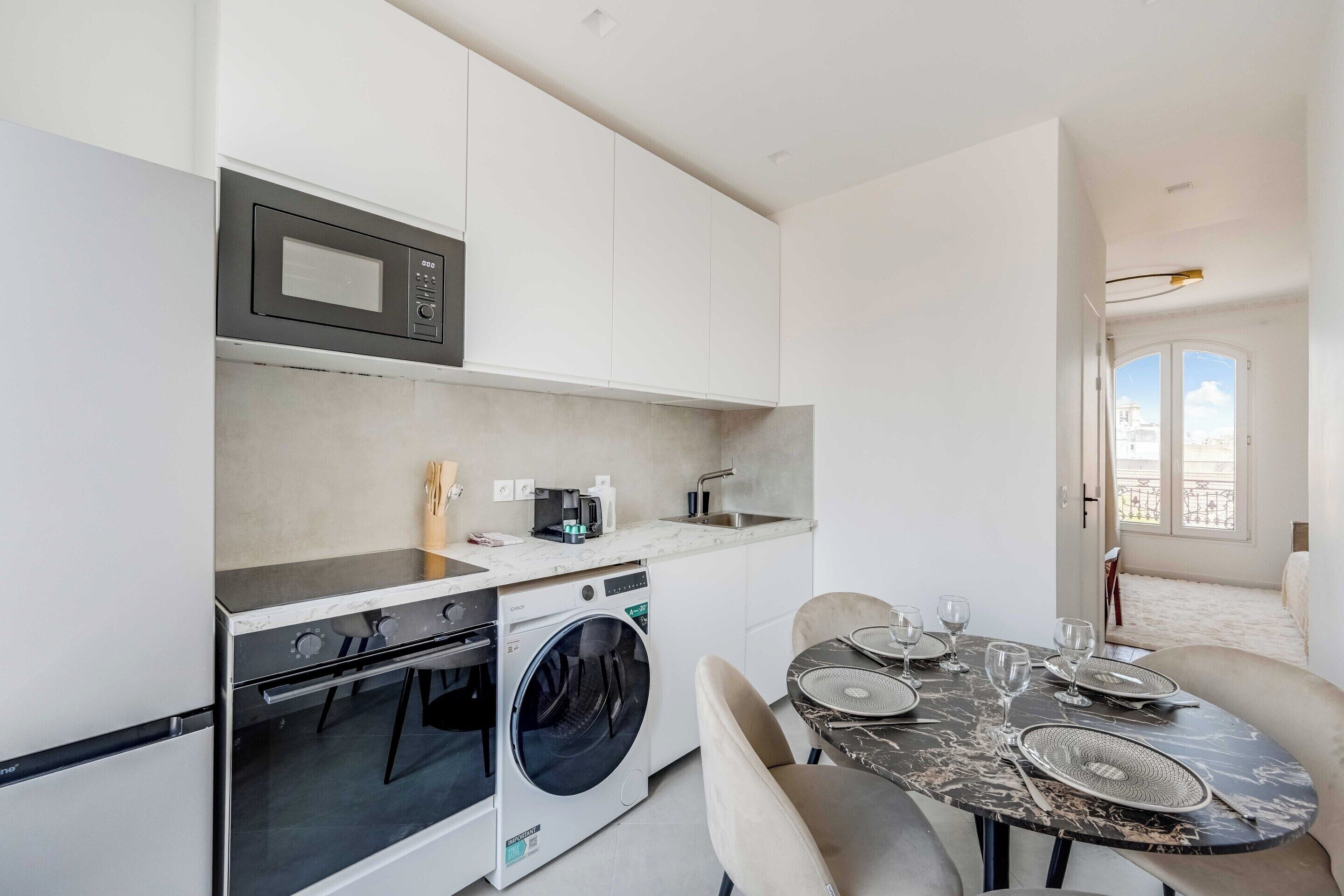 A compact kitchen with a microwave, oven, and washing machine, a dining table with chairs, and a view of the outside.