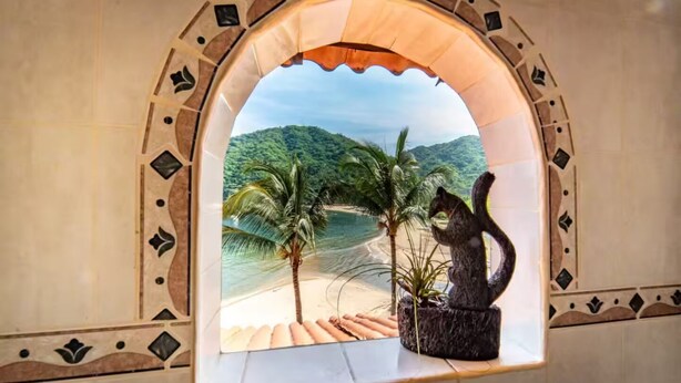 Une fenêtre donnant sur une plage et des palmiers, encadrée par une arche décorative.