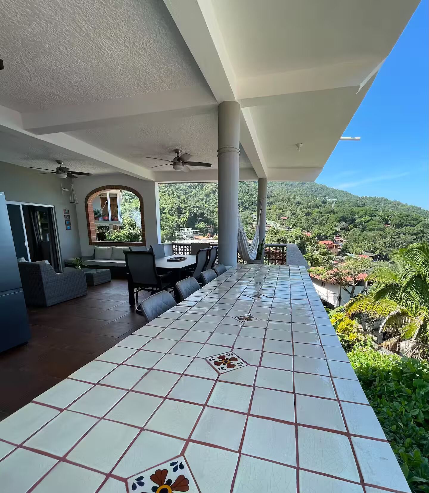 A covered terrace with a tiled floor, a dining area with chairs and a table, and a view of a lush green landscape.