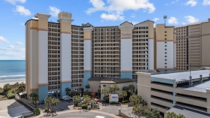 A multi-story hotel with a beach view, featuring a central courtyard with palm trees and a parking garage. - Luxury Beachfront Condo: King Suite in Premier Location in North Myrtle Beach (North Myrtle Beach)