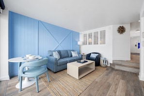 A modern living room with a blue accent wall, a sofa, a coffee table, and a dining area with a table and chairs. - Luxury Beachfront Condo: King Suite in Premier Location in North Myrtle Beach (North Myrtle Beach)
