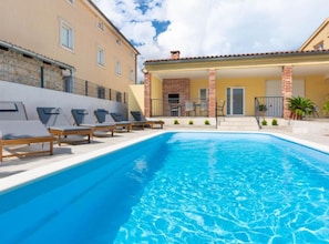 A swimming pool with lounge chairs, a covered patio area, and a building with a tiled roof.