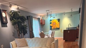 Una sala de estar con sofá, reposapiés y sillón de cuero, una mesa de comedor, una planta grande y una cocina con fregadero y armarios.