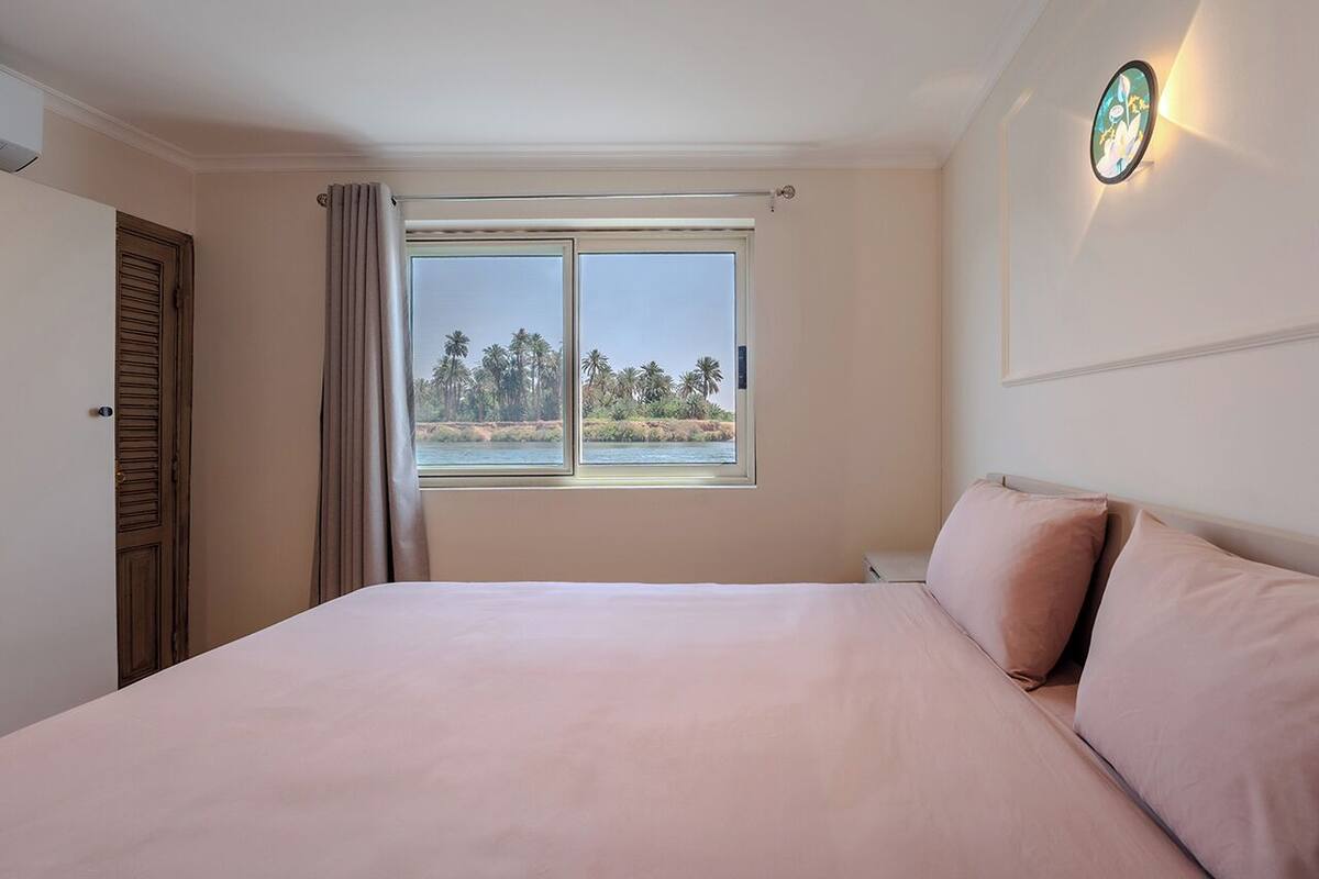 A bedroom with a large bed, a window with a view of palm trees, and a wall-mounted light fixture.