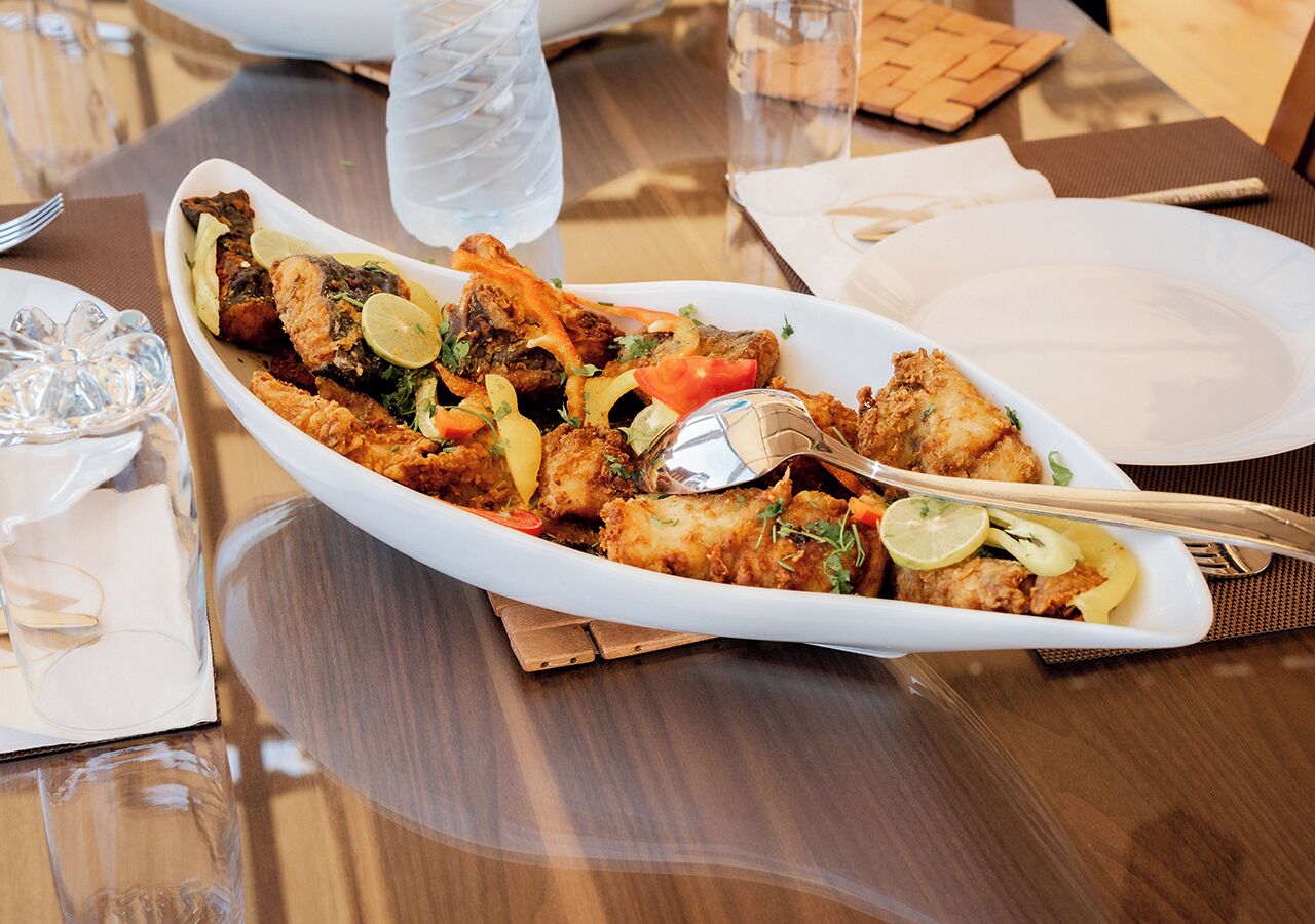 Un plat de fruits de mer servi dans une assiette ovale blanche, accompagné d’une cuillère, d’un verre d’eau et d’une serviette sur une table en bois.