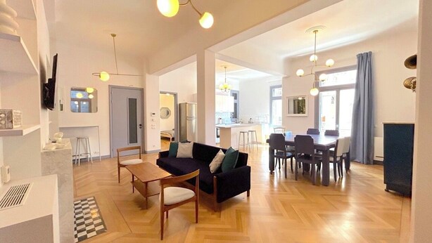 Amplia sala de estar con sofá, mesa de comedor y sillas, una cocina al fondo y un ventanal con cortinas.