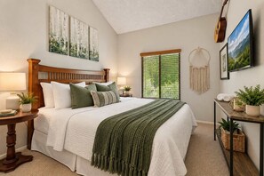 A bedroom with a wooden headboard, a large bed with white bedding, a green throw blanket, a bedside table with a lamp, and a window with blinds. - NEW | 18th Hole Retreat | Fam Friendly | Mtn Views (Flagstaff)