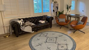 Een woonkamer met een leren bank, twee fauteuils, een rond tapijt, een boekenkast en een eettafel.