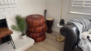 Een woonkamer met een witte bakstenen schoorsteen, een potplant, een stapel ronde ottomannen, een trommel, een lamp en een leren bank.