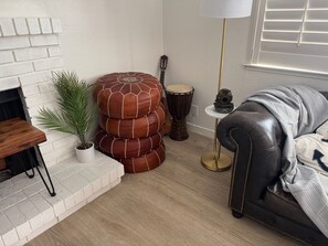 Ein Wohnzimmer mit Kamin aus weißen Ziegelsteinen, einem Blumentopf, einem Stapel runder Hocker, einer Trommel, einer Lampe und einem Ledersofa.