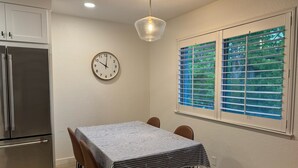 Een eethoek met een tafel, stoelen, een klok, een raam met jaloezieën en een koelkast.