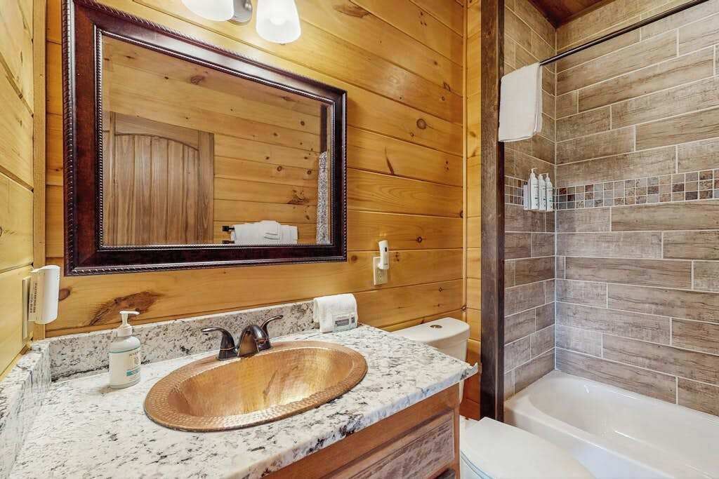 A wooden-paneled bathroom with a large mirror, a granite countertop, a copper sink, a toilet, and a shower area.