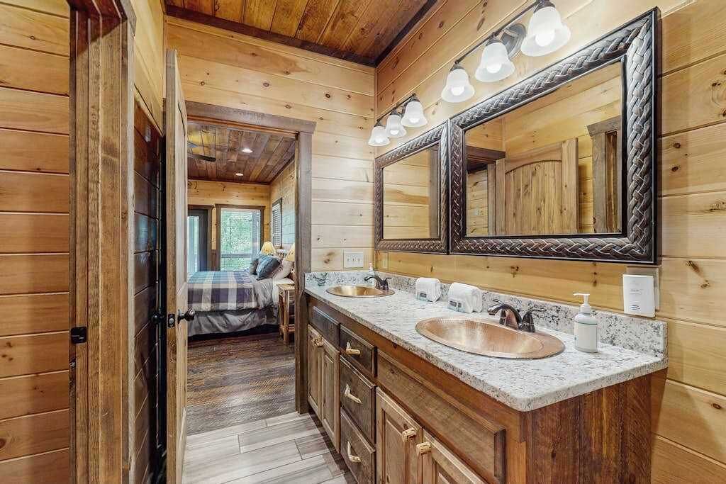 A wooden cabin interior with a large mirror, two sinks, and a bed visible through an open door.
