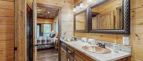 A wooden cabin interior with a large mirror, two sinks, and a bed visible through an open door.
