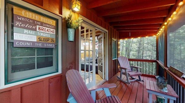 A wooden deck with Adirondack chairs, a rocking chair, and a table with a lantern. A sign with cabin rules is visible on the wall. - Lazy Days Cabin Stay (Blue Ridge)