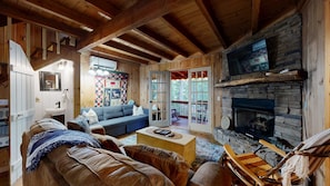 A cozy cabin interior with a stone fireplace, wooden beams, a blue sofa, and a view of the outdoors. - Lazy Days Cabin Stay (Blue Ridge)