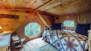 Un dormitorio tipo cabaña con techo de madera, una ventana grande, una cama con colcha estampada y una mesita pequeña.
