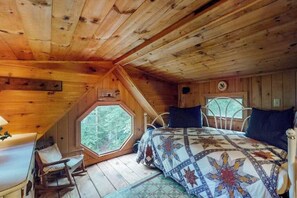 Ein Schlafzimmer einer Holzhütte mit Holzbalkendecke, einem großen Fenster, einem Bett mit gemustertem Quilten und einem kleinen Tisch.