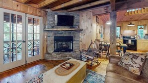 Interior rústico de una cabaña con chimenea de piedra, vigas de madera, comedor y cocina.