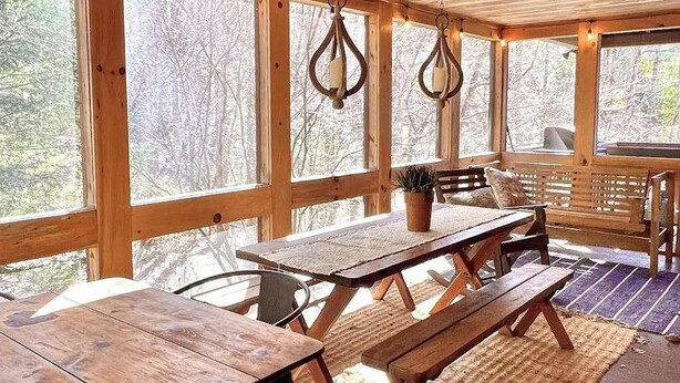 Eine Holzterrasse mit Picknicktisch und Bänken, großen Fenstern und hängenden Leuchten.