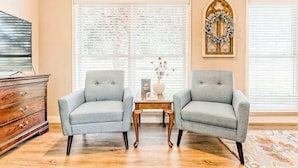 Un salon avec deux fauteuils gris, une commode en bois, une petite table sur laquelle est posé un vase, et une guirlande décorative accrochée au mur.