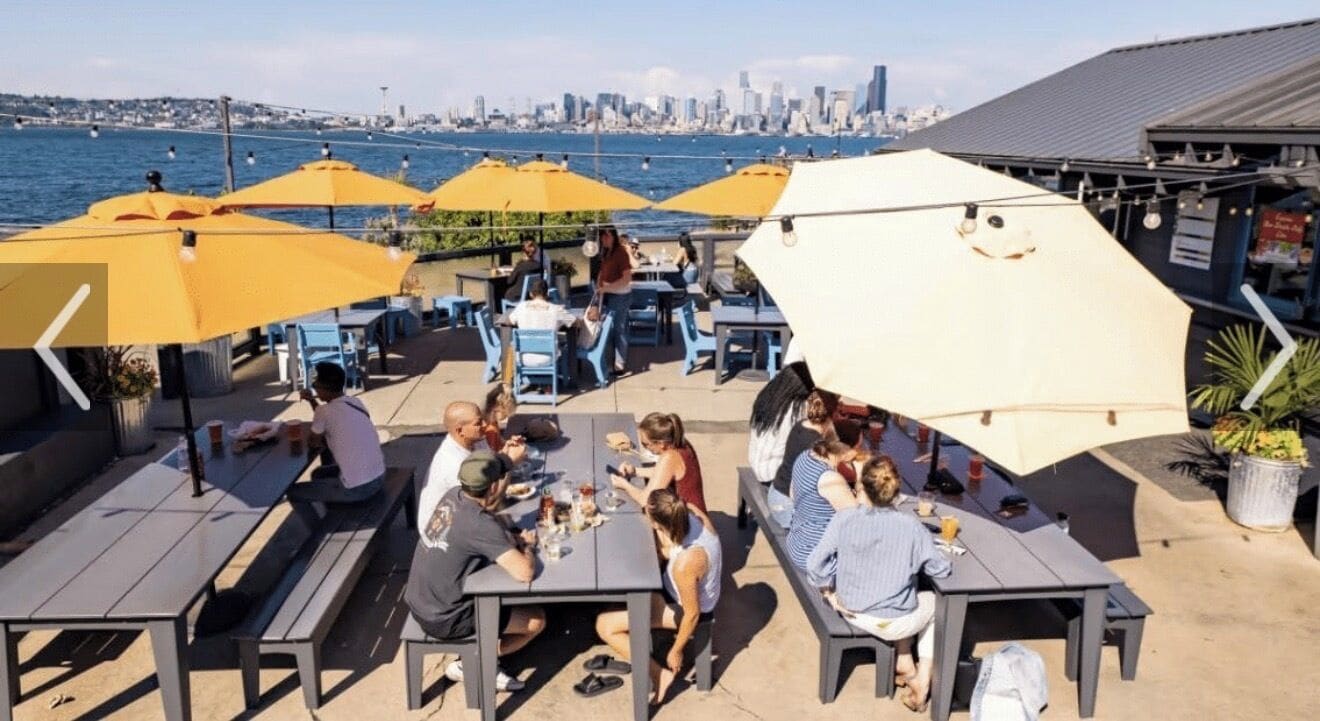 An outdoor dining area with tables and yellow umbrellas, overlooking a body of water and a city skyline.