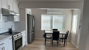 Et moderne køkken med rustfrit stål hvidevarer, et spiseområde med bord og stole, samt store vinduer med persienner.
