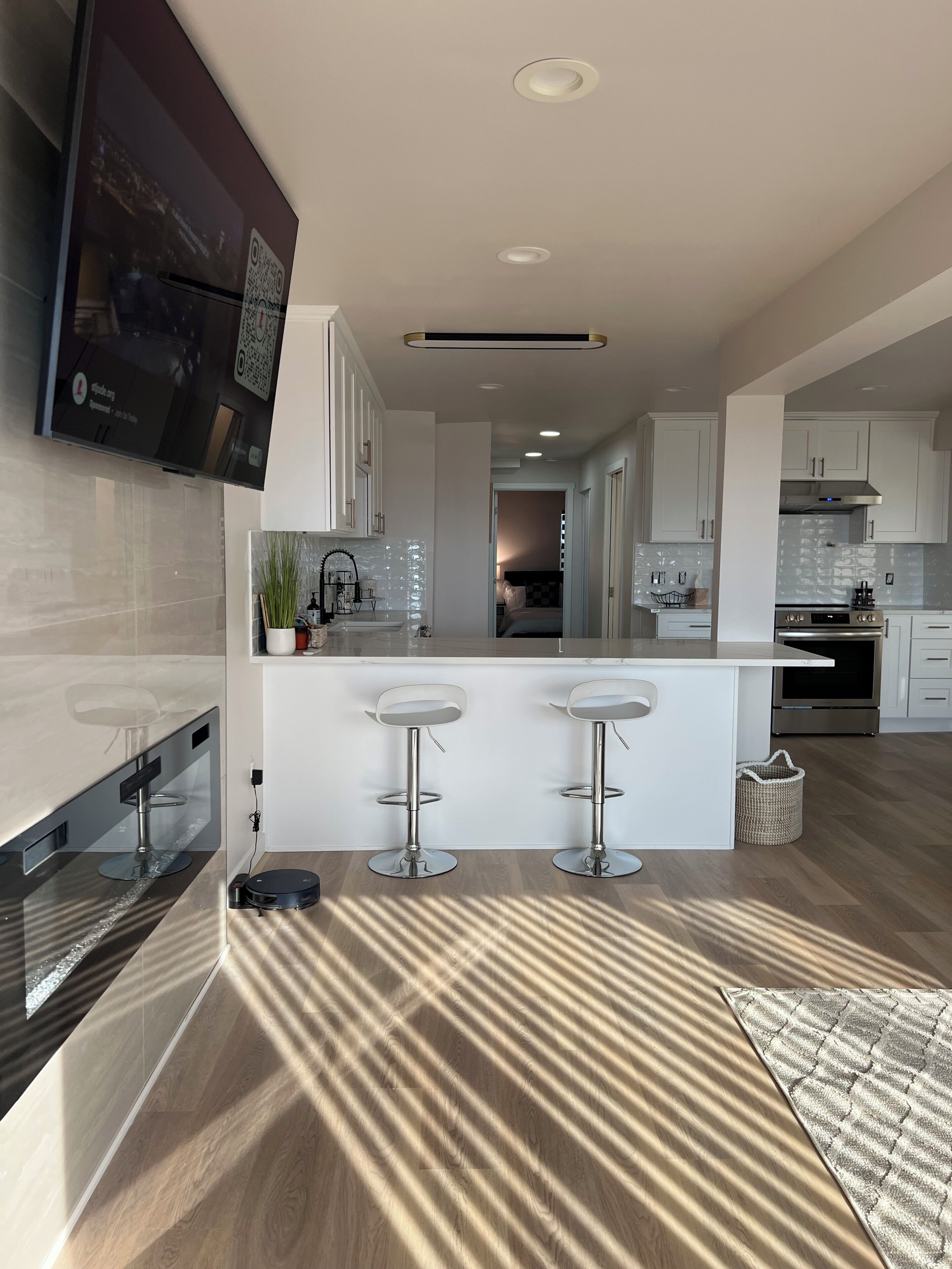 Una cocina moderna con un televisor montado en la pared, una isla central con taburetes de bar, un horno de acero inoxidable y un cubo de basura a la vista.