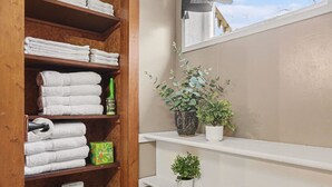 Een badkamer met een houten plankje met handdoeken en toiletpapier, een plant in pot en uitzicht op de lucht.