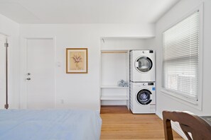 A bedroom with a bed, a washing machine, a wooden chair, and a window with blinds. - Charming Seaside Cottage in Victoria's Historic Core (Victoria South)