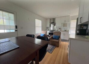 A modern kitchen with white cabinets, a stove, and a dining area with a table and chairs. - Charming Seaside Cottage in Victoria's Historic Core (Victoria South)