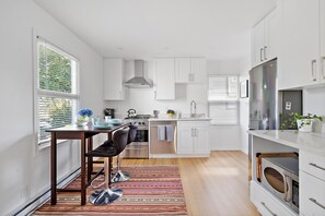 A modern kitchen with white cabinets, stainless steel appliances, and a dining area with a striped rug. - Charming Seaside Cottage in Victoria's Historic Core (Victoria South)
