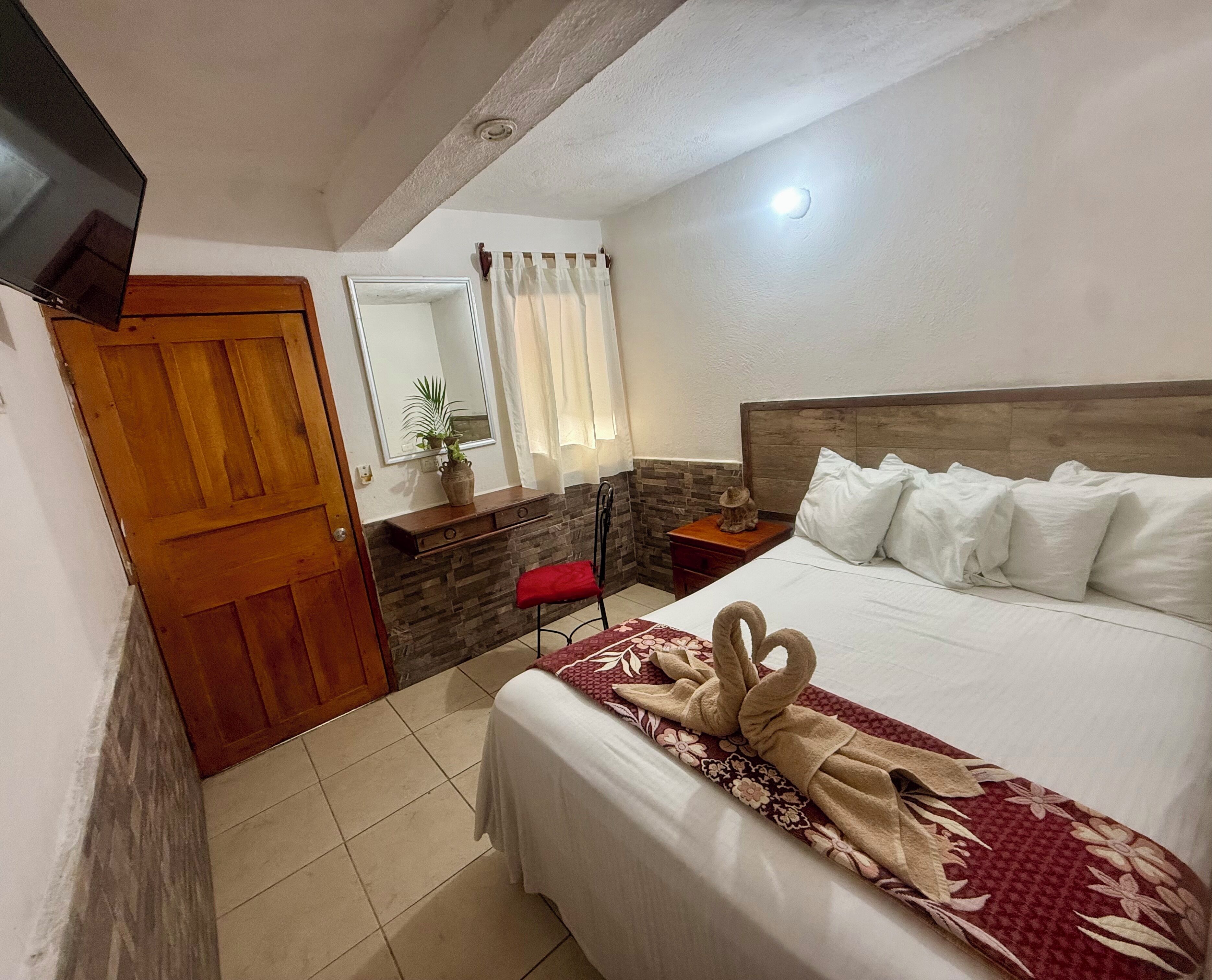 A hotel room with a wooden door, a mirror, a plant on a shelf, a bed with white pillows, and a red chair.