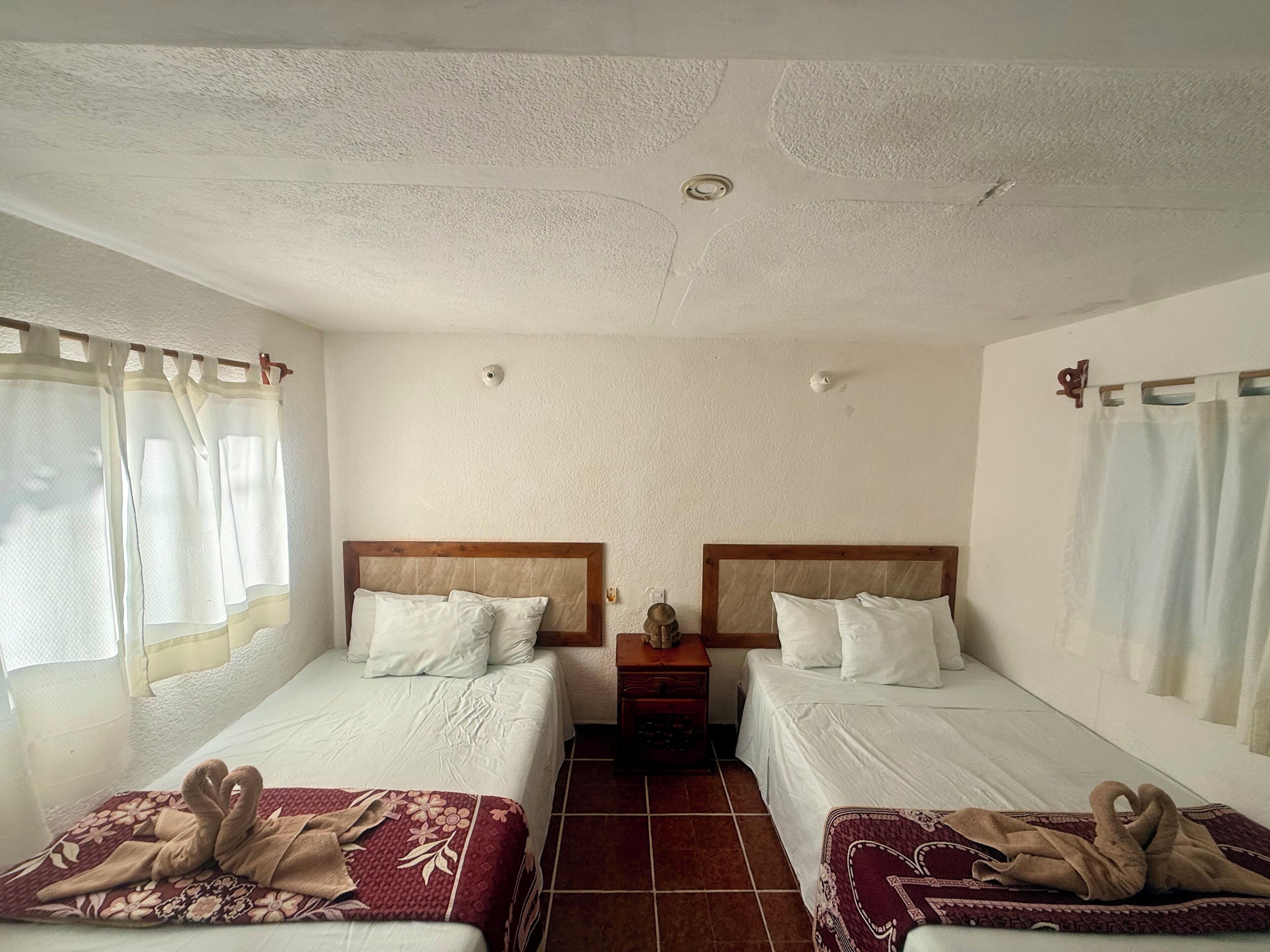 Habitación de hotel con dos camas, cabecero de madera, una mesita de noche y suelo de baldosas.