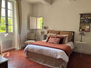 Une chambre à coucher avec un lit, une chaise, une table de chevet, une lampe et une fenêtre avec des rideaux.