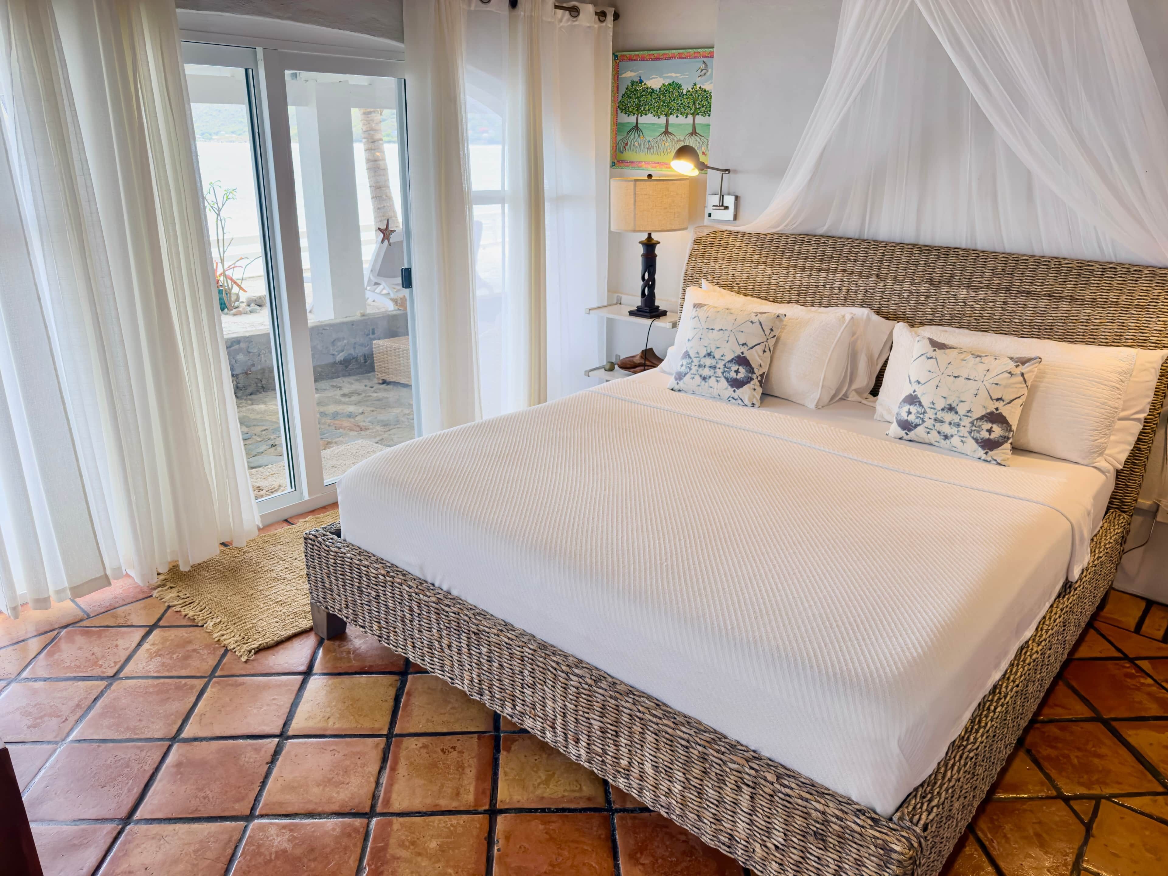 A bedroom with a wicker bed, white bedding, a nightstand with a lamp, and a view through a glass door.