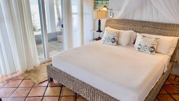 A bedroom with a wicker bed, white bedding, a nightstand with a lamp, and a view through a glass door.