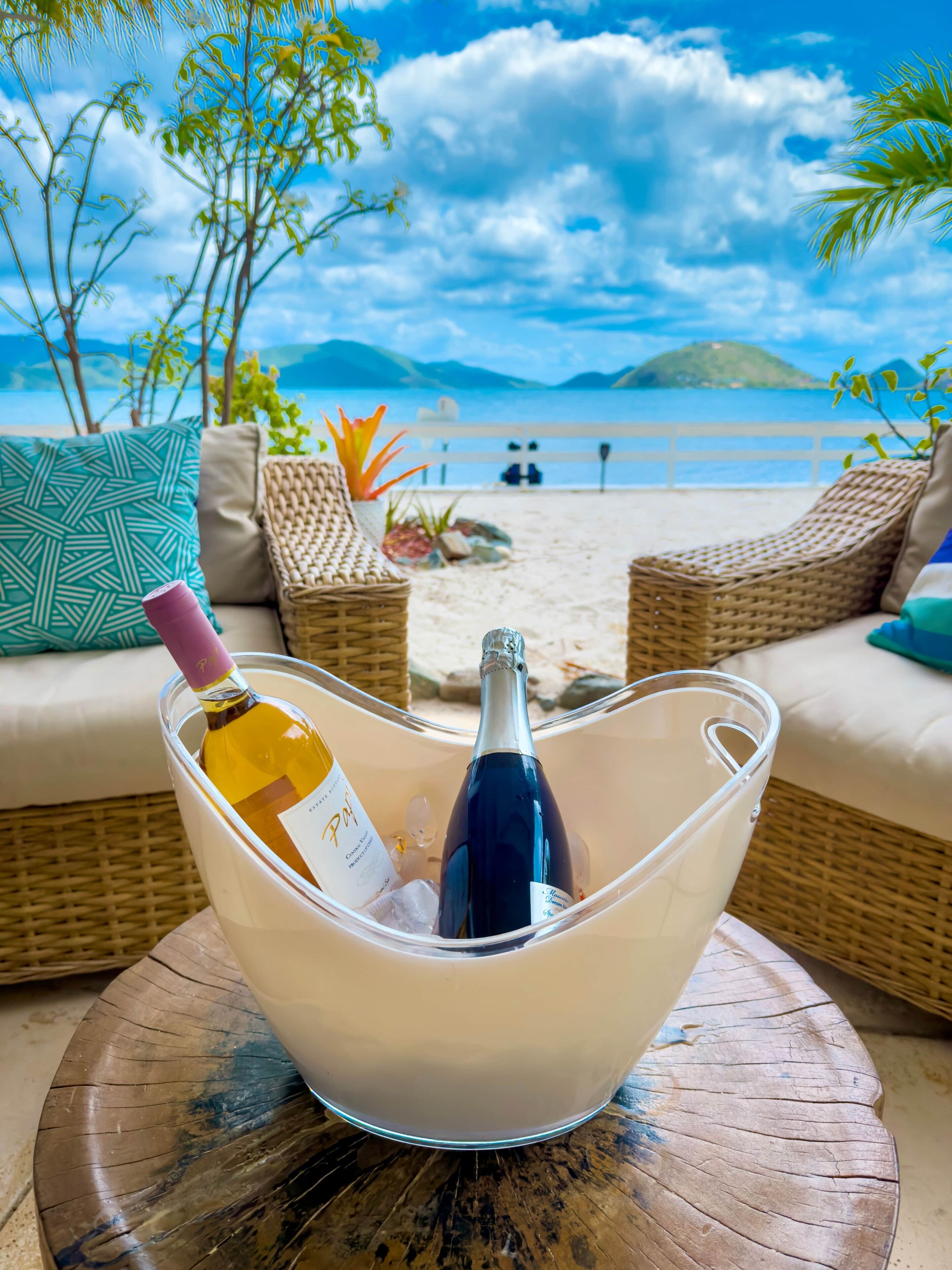 A beachside view with a wicker lounge chair, a cushioned sofa, and a clear glass ice bucket containing two bottles of wine.