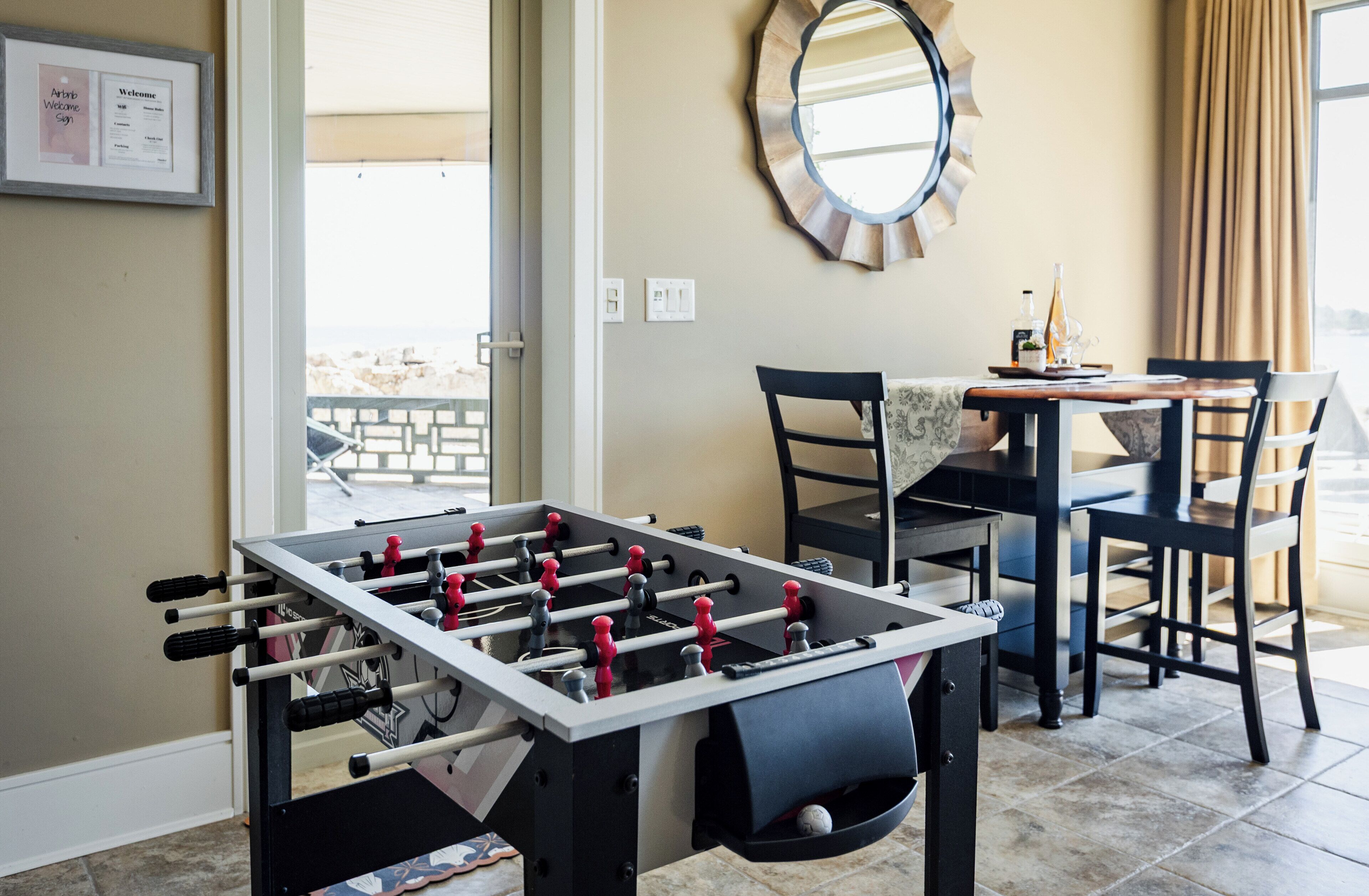 Une table de baby-foot dans une pièce qui contient également une table à manger, des chaises, un miroir rond et une fenêtre avec des rideaux.