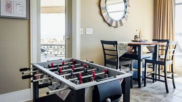 Une table de baby-foot dans une pièce qui contient également une table à manger, des chaises, un miroir rond et une fenêtre avec des rideaux.