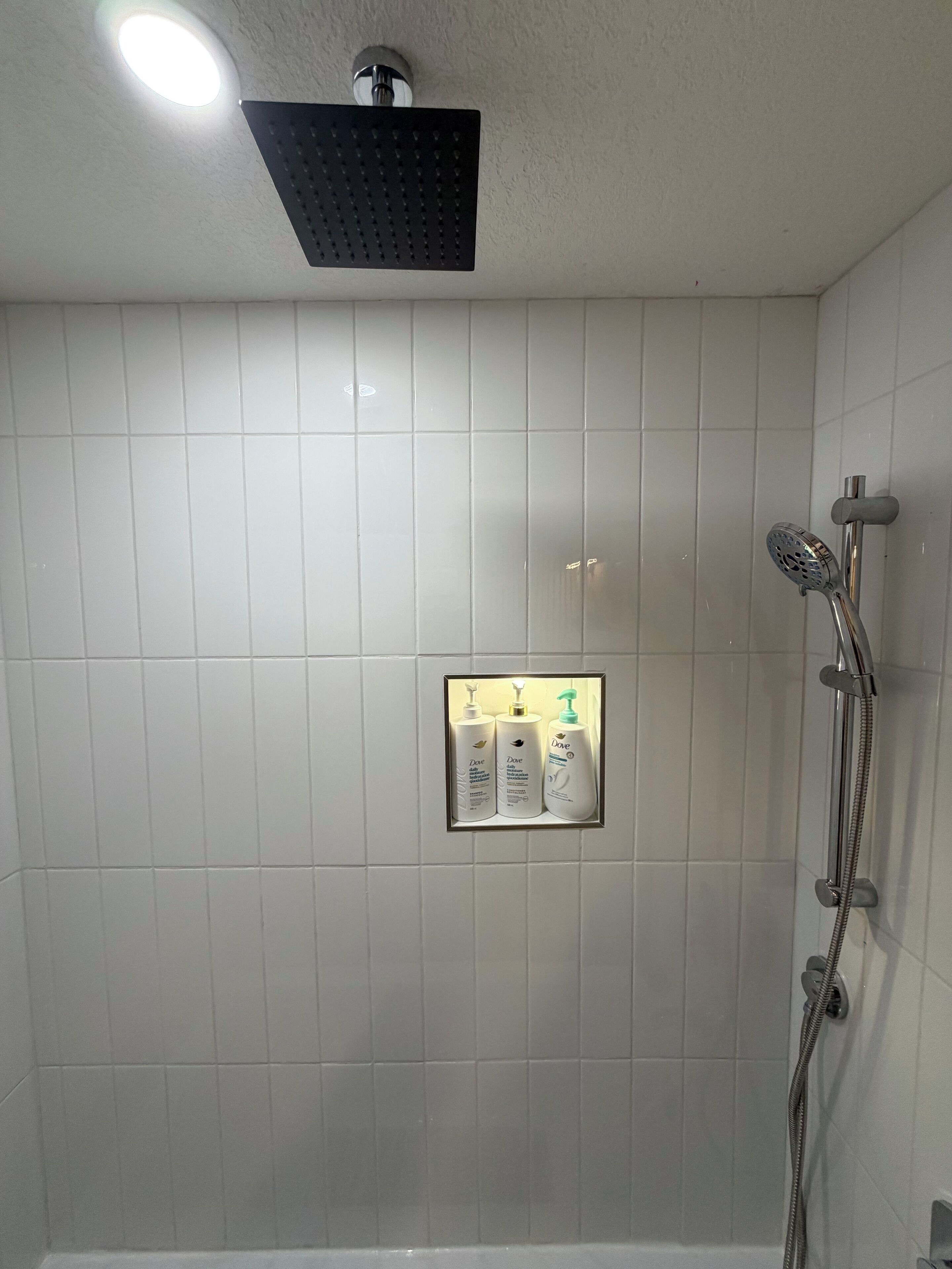 Un baño con azulejos blancos, ducha con cabezal de ducha de mano e iluminación empotrada en la pared.