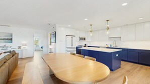 A modern kitchen with white cabinetry, a central island, and a dining area with a long table. - Rehoboth Beach - 24 Brooklyn Ave Unit E (Rehoboth Beach)