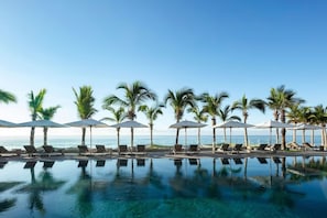 A poolside area with lounge chairs and umbrellas, palm trees, and a view of the ocean. - Christmas in Cabo! Garza Blanca Los Cabos – 2-Bedroom Luxury Suite (Cabo San Lucas)