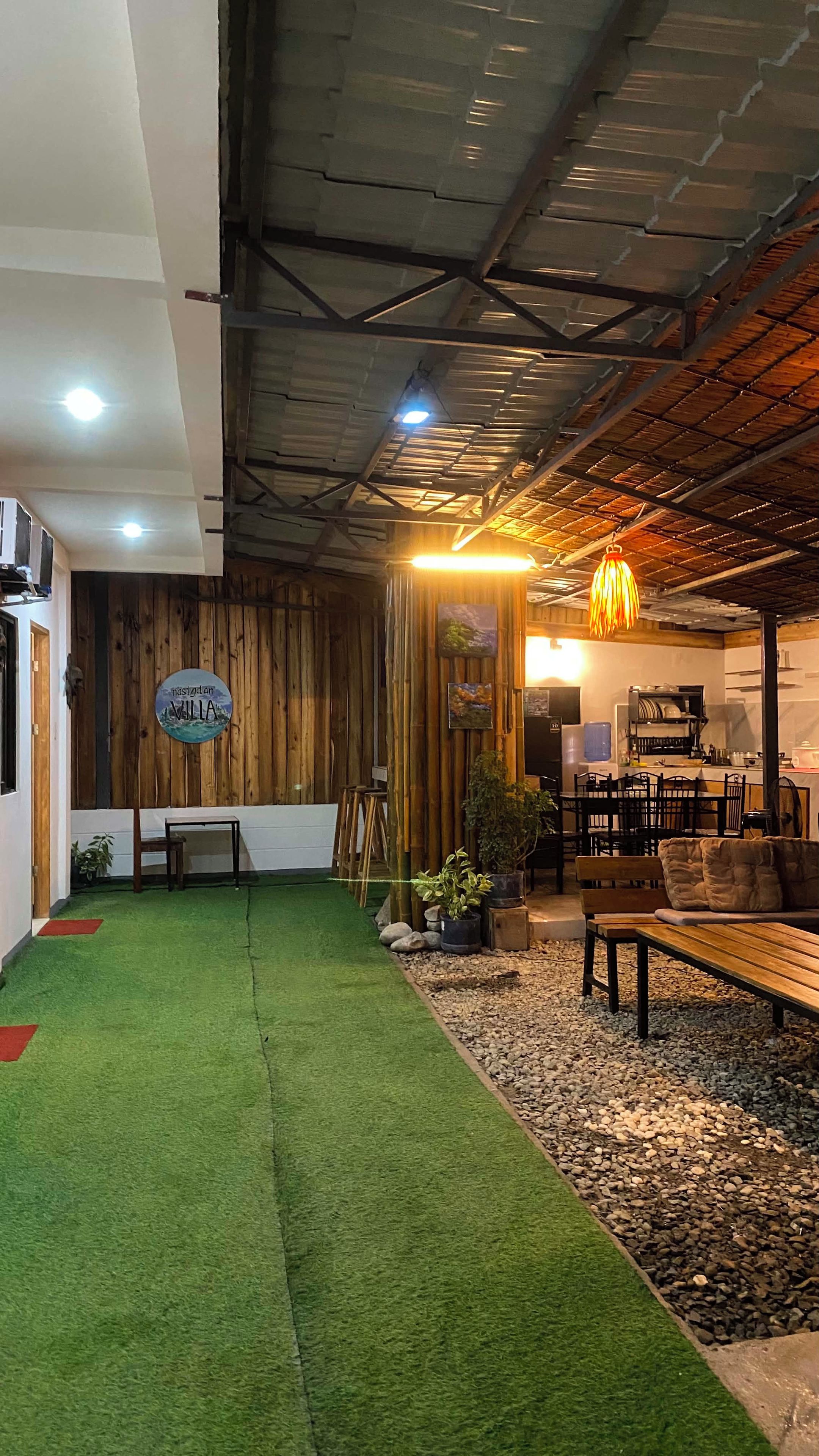 Un intérieur comprenant un espace recouvert d’un gazon synthétique vert, des murs en bois, un coin repas avec des tables et des chaises, et une cuisine à l’arrière-plan.