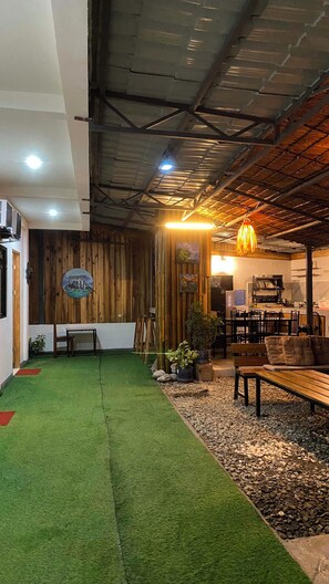 An interior with a green artificial turf area, wooden walls, a dining area with tables and chairs, and a kitchen in the background. - Charming 3-bedroom apartment with AC, smart tv , fridge ,wifi, kitchen ,& garage (Elnido)