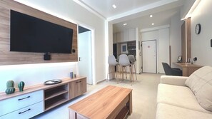 A modern living room with a flat-screen TV, wooden furniture, a dining area, and a kitchen in the background. - Modern Bright 1BR next to Pasalimani & Marina Zeas (Pireas)