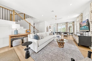 A modern living room with a white sofa, a coffee table, a staircase, and a dining area with a view of the outdoors. - Surfside 108 CRE (Port Aransas)