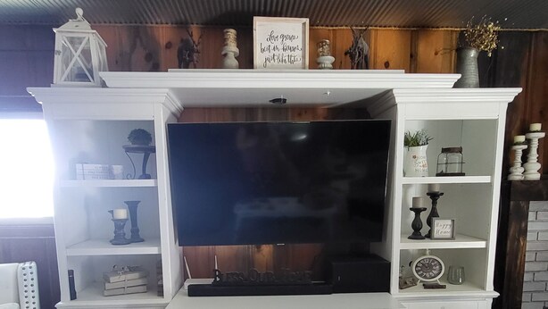 A white entertainment center with a flat-screen TV, shelves with decorative items, and a sign above the TV. - Breathtaking views surrounding this home, perfect for a relaxing getaway! (Cody)